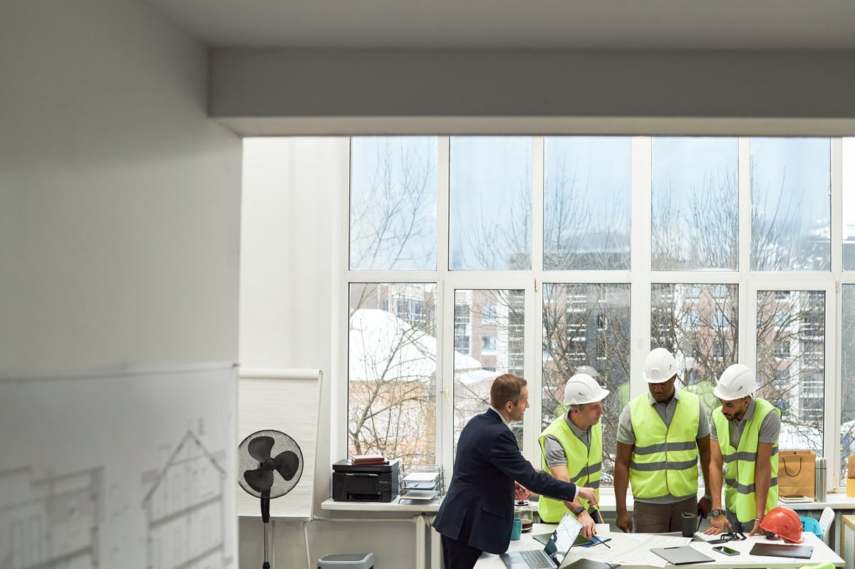 Caucasian middle aged man in suit discussing blueprints with two young adult men and one Black young adult man, wearing safety vests and hard hats, in office with large windows